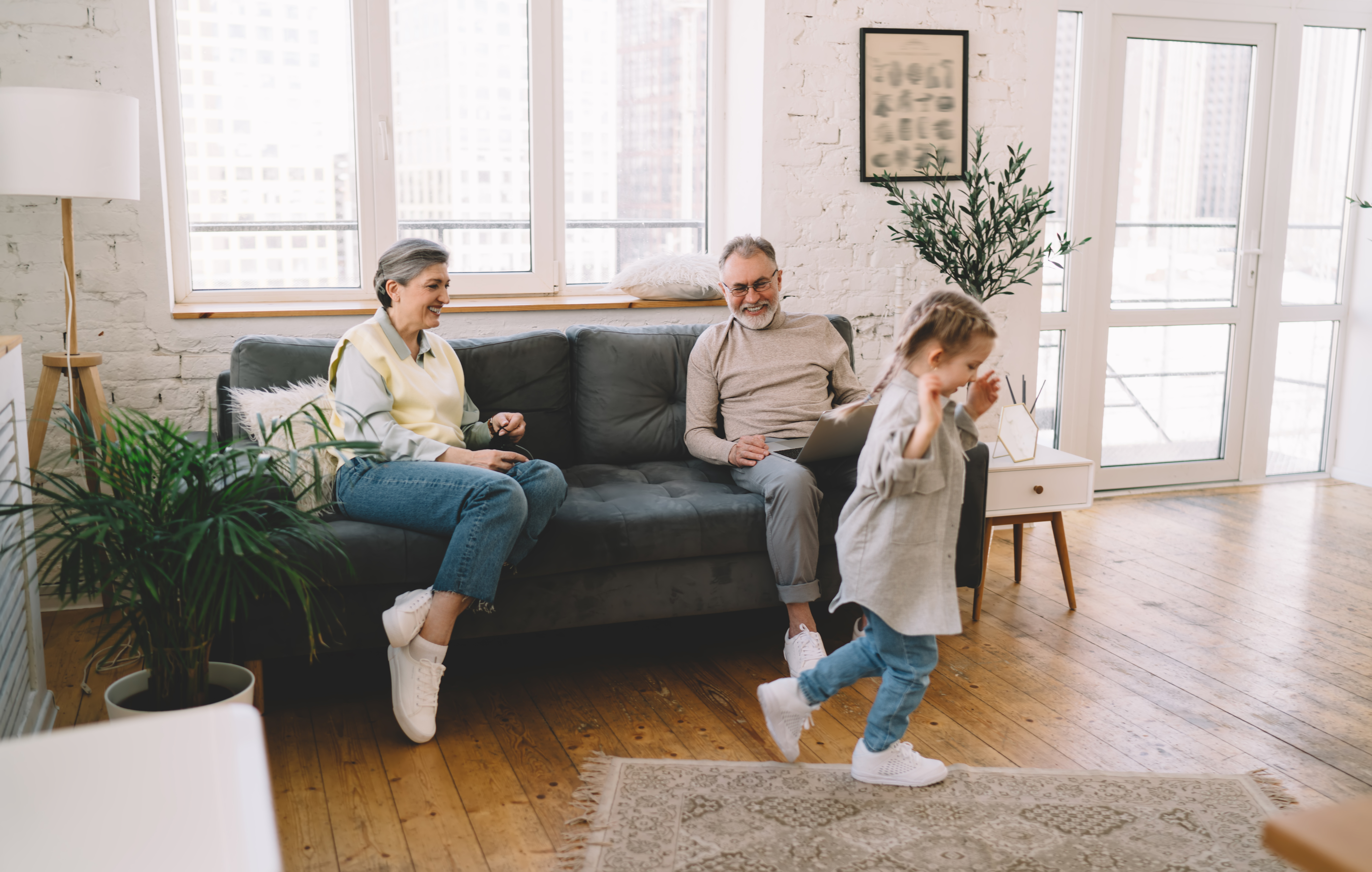 Full body of delighted elderly grandmother and grandfather looking at little granddaughter and smiling while sitting on comfortable couch in living room and relaxing on winter morning